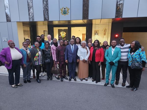 Group of smiling people, standing in front of a building. The are close together and look happy.